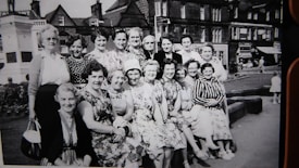 A group of women, dressed in vintage attire, pose together in an outdoor setting. They are seated and standing in front of a backdrop that includes a few buildings and a small park or square. The women are smiling, and the photograph appears to be black and white, suggesting an earlier time period. The area has a quaint, historical feel with visible architectural details.
