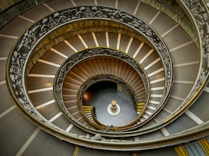 A spiral staircase captured from above, showcasing intricate metalwork on the railings and a central column at the base. The steps are broad and decorated with a pattern, creating a sense of depth and movement.