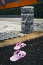 A pair of pink slippers lies on a wet urban surface near a stone bollard. The street and surrounding area show signs of recent rain, indicated by the pooled water. The background features wet pavement, a curb, and some greenery with a partial view of an orange structure.