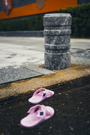 A pair of pink slippers lies on a wet urban surface near a stone bollard. The street and surrounding area show signs of recent rain, indicated by the pooled water. The background features wet pavement, a curb, and some greenery with a partial view of an orange structure.