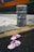 A pair of pink slippers lies on a wet urban surface near a stone bollard. The street and surrounding area show signs of recent rain, indicated by the pooled water. The background features wet pavement, a curb, and some greenery with a partial view of an orange structure.