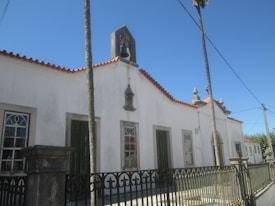 A white, historic-looking building features a red-tiled roof with decorative elements, including a small bell tower and sculpted details above the entrance. The structure is bordered by a wrought iron fence, and two tall palm trees stand in front of it. The building has arched windows with intricate designs.
