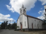 A white church with an aged exterior stands prominently against a backdrop of a blue sky with scattered clouds. The building features Gothic-style arched windows and a bell tower with a cross at the top. Surrounding the church are some trees and a clear, paved road.