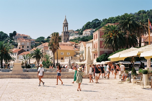 A scenic town square with people leisurely walking and exploring. The area is surrounded by Mediterranean architecture, featuring stone buildings with red-tiled roofs. A clock tower stands prominently in the background, while palm trees add a tropical flair. Outdoor cafes with yellow canopies and potted plants are scattered around, and the atmosphere feels vibrant and relaxed.