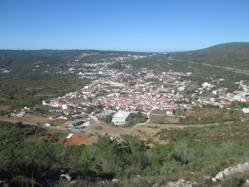 Aerial view of a small town nestled in a valley, surrounded by green hills and sparse vegetation. The town is characterized by clustered white and red-roofed buildings. There are visible roads that connect different parts of the town, and grassy areas can be seen surrounding the settlement.