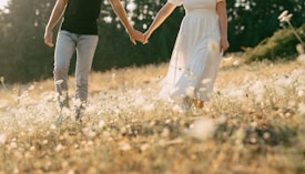 A couple walks hand in hand through a sunlit meadow filled with wildflowers. The woman wears a flowing white dress, and the man is in a black shirt and light jeans. Soft golden light filters through the trees in the background, creating a serene and romantic atmosphere.
