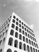 A striking, angular perspective of a large, modern architectural building featuring numerous arches. The structure appears to be made of light-colored stone with a repetitive pattern of arched windows spanning multiple floors. The sky is overcast, enhancing the monochromatic appearance of the image.
