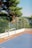 A small outdoor sports court with a blue surface and red borders, featuring a white and blue soccer goal. The court is surrounded by a net fence and overlooks a scenic view of lush green trees and a body of water in the background.