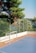 A small outdoor sports court with a blue surface and red borders, featuring a white and blue soccer goal. The court is surrounded by a net fence and overlooks a scenic view of lush green trees and a body of water in the background.