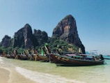 Several colorful longtail boats are anchored along a clear, sandy shoreline. Tall limestone cliffs rise impressively in the background under a bright blue sky, surrounded by lush green vegetation.