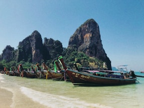 Several colorful longtail boats are anchored along a clear, sandy shoreline. Tall limestone cliffs rise impressively in the background under a bright blue sky, surrounded by lush green vegetation.