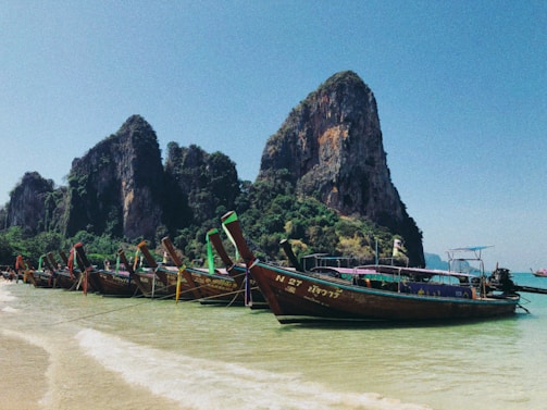 Several colorful longtail boats are anchored along a clear, sandy shoreline. Tall limestone cliffs rise impressively in the background under a bright blue sky, surrounded by lush green vegetation.