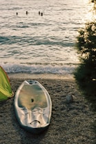 A stand-up paddleboard rests on a pebbly beach, with gentle waves lapping the shore. In the distance, three swimmers are visible in the water as the sun sets, casting a warm golden hue over the scene. A green tent is partially visible on the left side, and there's a bush with glistening leaves on the right.