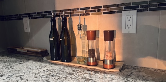 A kitchen countertop is neatly arranged with several bottles and grinders. Three dark glass bottles with metal spouts likely contain different oils or vinegars. Two transparent grinders with wooden tops are filled with peppercorns and Himalayan pink salt. The items are placed on a cream-colored tray, with a textured countertop and backsplash featuring a tile pattern.