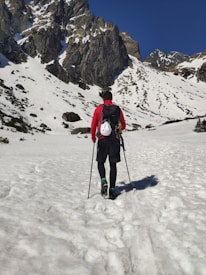 A person wearing a red jacket and black hiking gear is trekking through a snow-covered mountainous landscape. The mountains in the background are steep with rocky surfaces and partially covered in snow. The clear blue sky suggests a bright, sunny day, providing good visibility for the hike.