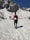 A person wearing a red jacket and black hiking gear is trekking through a snow-covered mountainous landscape. The mountains in the background are steep with rocky surfaces and partially covered in snow. The clear blue sky suggests a bright, sunny day, providing good visibility for the hike.