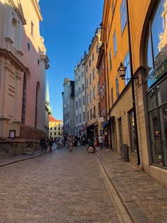A narrow cobblestone street lined with historic buildings of different colors in a picturesque European city. People are casually walking and exploring the area. The sky is clear with a bright blue hue, suggesting a pleasant weather.