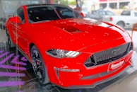 A vibrant red sports car is positioned indoors on a black reflective platform with purple lighting. The car's design is sleek and aerodynamic, highlighted by its prominent front grille and sharp headlights.