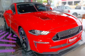 A vibrant red sports car is positioned indoors on a black reflective platform with purple lighting. The car's design is sleek and aerodynamic, highlighted by its prominent front grille and sharp headlights.