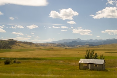 A wide expanse of open farmland is visible under a clear blue sky with scattered white clouds. Rolling hills stretch into the distance, with a range of mountains visible on the horizon. A small building with a corrugated metal roof stands in the foreground, surrounded by patches of green grass and a few sparse trees.