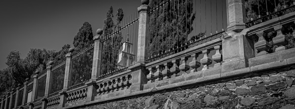 A black and white image featuring a decorative stone and wrought iron fence. The structure is composed of stone pillars with intricate ironwork in between, set against a background of tall cypress trees. The craftsmanship of the stone masonry and the design of the iron bars give the fence an elegant and historical appearance.