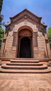 An ornate stone chapel with intricate carved details stands at the top of a series of steps. The building features decorative elements such as columns and crosses on the roof. The structure appears to be made of a reddish-brown stone, giving it an aged appearance. Trees flank the chapel, and a bright blue sky is visible above.