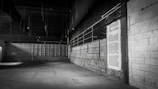 An empty, dimly lit warehouse interior with concrete walls and a high ceiling. The floor is spacious and devoid of any objects, and there are exposed pipes and metal beams visible. The color palette is muted, with shades of gray dominating the scene.