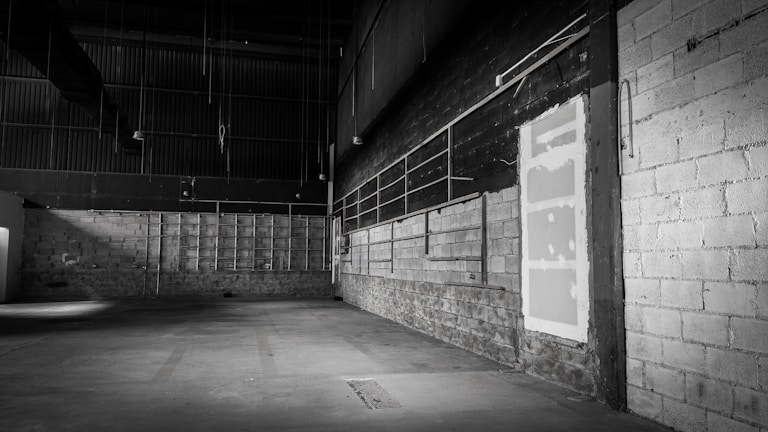 An empty, dimly lit warehouse interior with concrete walls and a high ceiling. The floor is spacious and devoid of any objects, and there are exposed pipes and metal beams visible. The color palette is muted, with shades of gray dominating the scene.