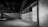 An empty, dimly lit warehouse interior with concrete walls and a high ceiling. The floor is spacious and devoid of any objects, and there are exposed pipes and metal beams visible. The color palette is muted, with shades of gray dominating the scene.