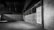 An empty, dimly lit warehouse interior with concrete walls and a high ceiling. The floor is spacious and devoid of any objects, and there are exposed pipes and metal beams visible. The color palette is muted, with shades of gray dominating the scene.