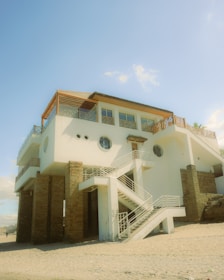 A modern beach house elevated on stilts stands on a sandy shore. The structure features multiple levels with white and brick facades. There are external staircases leading to different levels of the house, and it is topped with a sloped roof. The house includes several circular and square windows, and the surrounding sky is clear and blue.