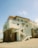 A modern beach house elevated on stilts stands on a sandy shore. The structure features multiple levels with white and brick facades. There are external staircases leading to different levels of the house, and it is topped with a sloped roof. The house includes several circular and square windows, and the surrounding sky is clear and blue.