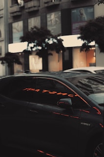 A sleek, dark-colored hybrid car parked on a city street reflects neon lights on its side windows. The background features urban elements like buildings with storefronts and trees, suggesting a bustling city environment.