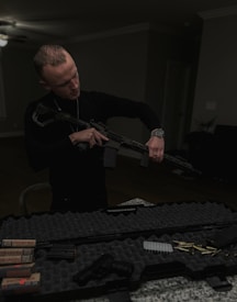 A man is standing indoors, holding a rifle while looking down at it. In front of him on a granite countertop is a hard-shelled gun case filled with various firearm accessories, including a handgun, several boxes of ammunition, and loose bullets.