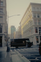 A street scene in an urban environment with a UPS delivery truck crossing the intersection. The surrounding buildings are made of pale stone with classical architecture. Street signs are visible, indicating the location is near Greene Street. The lighting suggests either early morning or late afternoon, casting a gentle glow on the buildings.