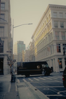 A street scene in an urban environment with a UPS delivery truck crossing the intersection. The surrounding buildings are made of pale stone with classical architecture. Street signs are visible, indicating the location is near Greene Street. The lighting suggests either early morning or late afternoon, casting a gentle glow on the buildings.