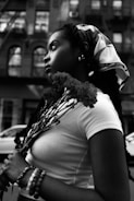 A black and white portrait of a person in profile, holding a bouquet of flowers. The individual has long braided hair and is wearing a headscarf and a white top. The urban background includes a building facade with windows and a parked car.