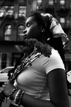 A black and white portrait of a person in profile, holding a bouquet of flowers. The individual has long braided hair and is wearing a headscarf and a white top. The urban background includes a building facade with windows and a parked car.
