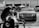 A luxury car is parked in an industrial garage setting. The car is sleek and shiny, with droplets of water on its surface, suggesting it has recently been washed. In the foreground, a blurry figure appears to be working, possibly detailing or inspecting another vehicle. Various car parts and tools are visible, along with other vehicles and equipment in the background.