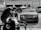 A luxury car is parked in an industrial garage setting. The car is sleek and shiny, with droplets of water on its surface, suggesting it has recently been washed. In the foreground, a blurry figure appears to be working, possibly detailing or inspecting another vehicle. Various car parts and tools are visible, along with other vehicles and equipment in the background.