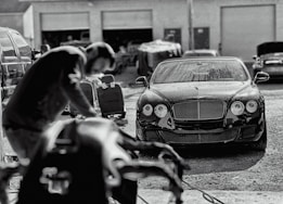 A luxury car is parked in an industrial garage setting. The car is sleek and shiny, with droplets of water on its surface, suggesting it has recently been washed. In the foreground, a blurry figure appears to be working, possibly detailing or inspecting another vehicle. Various car parts and tools are visible, along with other vehicles and equipment in the background.