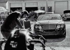 A luxury car is parked in an industrial garage setting. The car is sleek and shiny, with droplets of water on its surface, suggesting it has recently been washed. In the foreground, a blurry figure appears to be working, possibly detailing or inspecting another vehicle. Various car parts and tools are visible, along with other vehicles and equipment in the background.