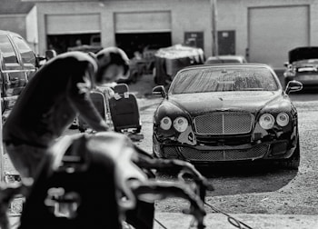 A luxury car is parked in an industrial garage setting. The car is sleek and shiny, with droplets of water on its surface, suggesting it has recently been washed. In the foreground, a blurry figure appears to be working, possibly detailing or inspecting another vehicle. Various car parts and tools are visible, along with other vehicles and equipment in the background.