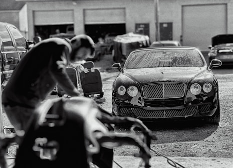 A luxury car is parked in an industrial garage setting. The car is sleek and shiny, with droplets of water on its surface, suggesting it has recently been washed. In the foreground, a blurry figure appears to be working, possibly detailing or inspecting another vehicle. Various car parts and tools are visible, along with other vehicles and equipment in the background.