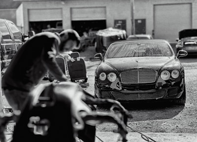A luxury car is parked in an industrial garage setting. The car is sleek and shiny, with droplets of water on its surface, suggesting it has recently been washed. In the foreground, a blurry figure appears to be working, possibly detailing or inspecting another vehicle. Various car parts and tools are visible, along with other vehicles and equipment in the background.