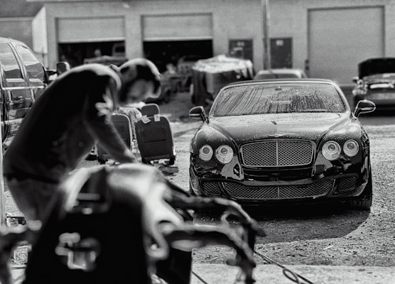 A luxury car is parked in an industrial garage setting. The car is sleek and shiny, with droplets of water on its surface, suggesting it has recently been washed. In the foreground, a blurry figure appears to be working, possibly detailing or inspecting another vehicle. Various car parts and tools are visible, along with other vehicles and equipment in the background.