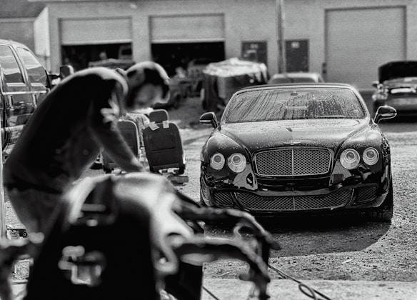 A luxury car is parked in an industrial garage setting. The car is sleek and shiny, with droplets of water on its surface, suggesting it has recently been washed. In the foreground, a blurry figure appears to be working, possibly detailing or inspecting another vehicle. Various car parts and tools are visible, along with other vehicles and equipment in the background.