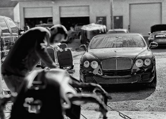 A luxury car is parked in an industrial garage setting. The car is sleek and shiny, with droplets of water on its surface, suggesting it has recently been washed. In the foreground, a blurry figure appears to be working, possibly detailing or inspecting another vehicle. Various car parts and tools are visible, along with other vehicles and equipment in the background.