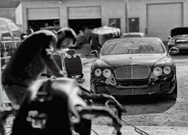 A luxury car is parked in an industrial garage setting. The car is sleek and shiny, with droplets of water on its surface, suggesting it has recently been washed. In the foreground, a blurry figure appears to be working, possibly detailing or inspecting another vehicle. Various car parts and tools are visible, along with other vehicles and equipment in the background.
