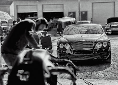 A luxury car is parked in an industrial garage setting. The car is sleek and shiny, with droplets of water on its surface, suggesting it has recently been washed. In the foreground, a blurry figure appears to be working, possibly detailing or inspecting another vehicle. Various car parts and tools are visible, along with other vehicles and equipment in the background.